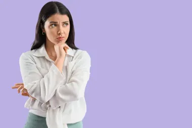 Zamyślona kobieta w białej koszuli spogląda w bok, z dłonią pod brodą na fioletowym tle.