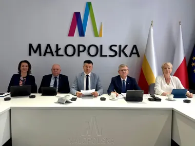 Pięć osób siedzi za białym stołem konferencyjnym z logo Małopolski na ścianie oraz flagami w tle, podczas spotkania lub konferencji prasowej.
