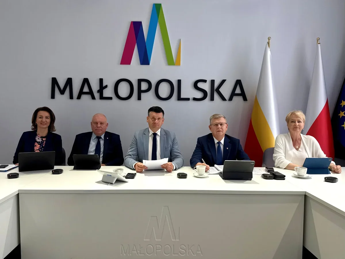Pięć osób siedzi za białym stołem konferencyjnym z logo Małopolski na ścianie oraz flagami w tle, podczas spotkania lub konferencji prasowej.